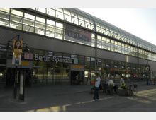 Außenansicht des Bahnhofs Berlin-Spandau mit einer modernen Glasfassade und einem gewölbten Dach. Vor dem Gebäude befinden sich Passanten, einige Fahrräder und ein kleiner Pflanzkübel.