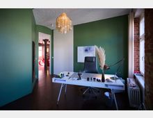 Arbeitszimmer mit grün gestrichener Wand, einem weißen Schreibtisch und einem schwarzen Bürostuhl. Im Raum sind eine große Fensterfront mit Ziegelwand, eine moderne Hängelampe und dekorative Trockenblumen sichtbar; im Hintergrund öffnet sich eine Tür zu einem farbenfrohen Raum mit Holzbalken.