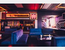 Innenraum einer modernen Lounge mit dunkler Einrichtung, blauen Polsterbänken und schwarzen Tischen. Neonbeleuchtung in warmen Farben, geometrische Wandmuster und Spiegel sowie eine Discokugel sorgen für ein futuristisches Ambiente.
