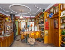 Ein historisches Lebensmittelgeschäft mit Holzmöbeln und Regalen, die gefüllt sind mit Glasflaschen, Behältern und Vintage-Waren. In der Mitte steht ein gusseiserner Ofen, und auf der Verkaufstheke befinden sich dekorative und funktionale Elemente wie alte Dosen und Reklameschilder.
