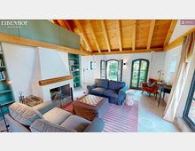 Gemütlicher Wohnraum mit sichtbarer Holzdecke, einem zentralen Kamin in weißer Wand, zwei grauen Sofas und einem Schachbrett auf einem Hocker. Große Fenster mit schwarzen Rahmen bieten Blick auf einen Garten, ergänzt durch Bücherregale und helle Vorhänge.