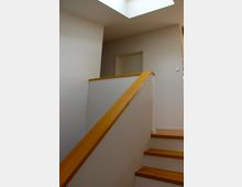 Treppe in einem modernen Innenraum mit Holzstufen und geländerähnlichem Handlauf aus Holz, weiß gestrichenen Wänden und einer rechteckigen Deckenöffnung, die natürliches Licht hereinlässt.