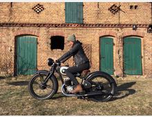 Eine Person auf einem klassischen Motorrad vor der Fassade eines alten Ziegelgebäudes mit grünen Holztüren und Fenstern. Die Szene befindet sich auf einer grasbedeckten Fläche bei Tageslicht.