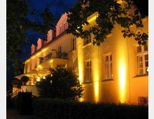 Beleuchtete Fassade eines zweistöckigen, historischen Gebäudes mit gelblichem Anstrich, großen Fenstern und einem Balkon in der Mitte. Vor dem Gebäude befinden sich Bäume und eine Terrasse mit Sonnenschirmen, aufgenommen bei Nacht.