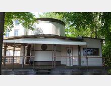 Rundes Gebäude mit modernem Design, Holzveranda und Flachdach, von Bäumen umgeben. Die Fassade ist weiß mit runden Fenstern, und im Hintergrund ist ein weiteres, größeres Gebäude sichtbar.