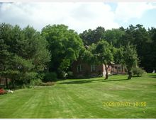 Backsteinhaus mit großen Fenstern, umgeben von einer weitläufigen, gepflegten Rasenfläche und Bäumen. Im Hintergrund dichter Wald, blauer Himmel und einige Wolken.
