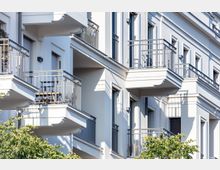 Mehrfamilienhaus mit weißer Fassade und mehreren Balkonen mit Metallgeländern. Im Vordergrund sind Bäume sichtbar, die teilweise die Balkone verdecken.