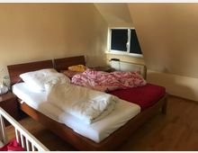 Doppelbett in einem schlichten Schlafzimmer mit Dachschräge, hellem Holzboden und cremefarbenen Wänden. Ein kleines Fenster mit Verdunkelungsschäden sowie ein Heizkörper befinden sich an der Wand nahe dem Bett.