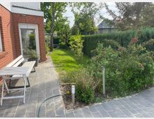 Gepflasterte Terrasse neben einem Backsteinhaus mit einer weißen Bank und einem Tisch, umgeben von einem gepflegten Garten mit Sträuchern, blühenden Pflanzen und einer Hecke im Hintergrund. Ein Gartenweg führt zu einer Rasenfläche, eingerahmt von Bäumen und weiteren Sträuchern.