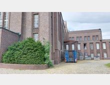 Backsteinbau mit großen Fenstern im industriellen Stil, gepflasterter Vorplatz und ein blauer Container neben mehreren Mülltonnen. Links im Vordergrund ist ein großer Busch in einem gemauerten Beet.