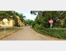 Gepflasterte Straße in einer Wohnsiedlung mit gelb gestrichenen Gebäuden auf der linken Seite und dichter Vegetation rechts. Im Vordergrund stehen ein rundes Verbotsschild und eine Straßenlaterne.