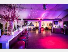 Eine Lounge mit Bar in einem Raum mit weißer, stoffbespannter Decke und farbiger Beleuchtung. Die Bar hat rote Hocker und eine Dekoration aus Zweigen, während im Hintergrund Sitzgruppen mit Sofas und Sesseln an Fenstern mit weißen Vorhängen stehen.