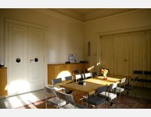 Heller Besprechungsraum mit einem großen Holztisch und mehreren Stühlen, umgeben von weißen Doppeltüren und schlichten Wänden. Im Hintergrund stehen hölzerne Schränke, auf denen eine Vase mit Tulpen und ein gerahmtes Dokument zu sehen sind.