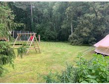 Wiese mit einem Spielplatz, der aus einer Holzschaukel mit Rutsche besteht, daneben ein Trampolin mit Netz. Im Hintergrund steht ein dichter Wald, rechts ragt ein Teil eines großen Zeltes ins Bild.