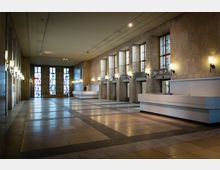 Großer Innenraum eines historischen Gebäudes mit hohen Decken, rechteckigen Steinsäulen, eleganten Wandlampen und großen, bunten Glasfenstern im Hintergrund. Rechts befindet sich eine moderne Empfangstheke, und über mehreren Türen hängen Beschilderungen mit Raumnummern.