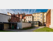 Hinterhof mit mehreren Gebäuden, darunter ein gelbes vierstöckiges Wohngebäude, ein rotes Backsteingebäude und weitere Nebengebäude. Pflasterwege sowie kleine Rasenflächen umgeben die Gebäude, und der Blick zeigt eine Mischung aus moderner und historischer Architektur.