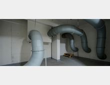 Ein technischer Raum mit großen grauen Lüftungsrohren, die geschwungen an der Decke und den Wänden entlanglaufen. Der Raum hat Betonwände, einen grauen Boden und eine hölzerne Palette in einer Ecke.