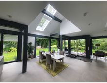 Innenraum eines modernen Wohnbereichs mit großen Glasfronten, die einen Blick auf einen grünen Garten bieten. Der Raum enthält eine Essecke mit weißem Tisch und grauen Stühlen, eine hohe Decke mit Dachfenstern und eine Terrasse, die durch Schiebetüren zugänglich ist.