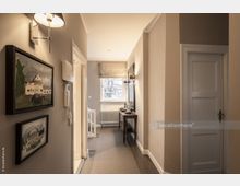 Schmaler, hell beleuchteter Flur in einem Wohnhaus mit beigen Wänden, einem Teppichläufer und Wanddekoration in Form von Bildern. Links befindet sich eine Tür und ein Treppenaufgang, rechts ein schmaler Holztisch unter einer Wandlampe vor einem Fenster mit Rollladen.