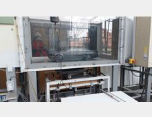 Ein Windkanal mit einem Modellauto hinter einer Glasscheibe, umgeben von technischen Geräten und Kabeln. Im Hintergrund sind ein Fenster und ein rotes Backsteingebäude zu sehen.