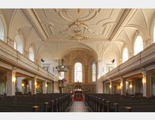 Innenraum einer historischen Kirche mit hellem barockem Dekor. Zu sehen sind eine verzierte Holzkanzel, hohe Rundbogenfenster, eine reich verzierte Decke und lange Sitzbänke, die zum Altar führen.