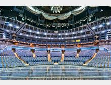 Innenansicht eines modernen Theaters mit mehreren Sitzreihen, die halbkreisförmig um die Bühne angeordnet sind. Die Architektur umfasst eine hohe Decke mit Bühnenbeleuchtung und technischem Equipment, sowie einen großen, offenen Saal mit blau gepolsterten Sitzen und Holzdetails an den Treppen.