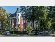 Straßenszene mit einer Reihe von historischen Wohnhäusern aus rotem Backstein und weißen Putzfassaden, darunter ein Gebäude mit einem kleinen Turm und spitzem Dach. Die Straße ist von hohen Bäumen gesäumt, mit parkenden Autos auf beiden Seiten, und eine Person geht auf dem Gehweg entlang.