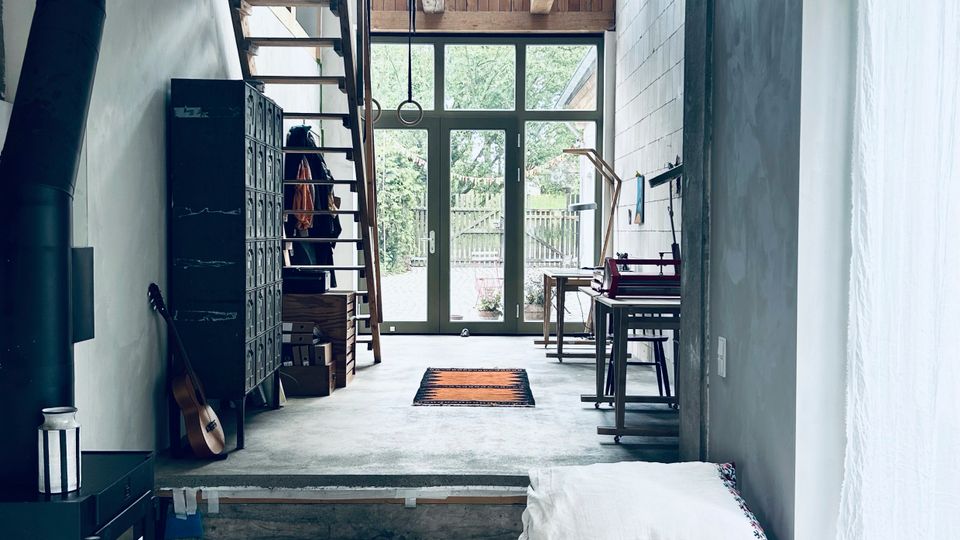 Innenraum eines modernen, loftartigen Wohnhauses mit Betonboden, Holzdeckenbalken und einem großen Oberlicht über dem Durchgang. Im Hintergrund führen eine offene Treppe und hohe Glastüren nach draußen; entlang der Wände stehen ein schwarzer Ofen, ein Spind, ein Arbeitsplatz sowie eine Liege mit gemustertem Teppich neben einem großen Fenster mit Vorhang.