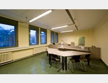 Ein schlicht eingerichteter Besprechungsraum mit hellgelben Wänden, mehreren Fenstern mit Blick nach draußen, einem angedeuteten Tresenbereich mit Bürostühlen sowie einem großen, hellen Besprechungstisch und dazugehörigen Stühlen. Der Raum besitzt grünen Bodenbelag und wird von Deckenleuchten beleuchtet.