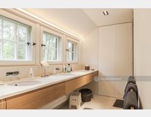 Helles Badezimmer mit langer Waschtischplatte aus Holz und zwei eingelassenen Waschbecken, darüber drei Fenster mit Sprossen, die den Blick ins Freie ermöglichen. An der rechten Wand befindet sich ein großer, weißer Einbauschrank, und Handtücher hängen auf einer Stange.