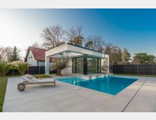 Moderner Poolbereich im Garten mit rechteckigem Swimmingpool, großer heller Terrassenfläche und einer minimalistischen Pavillonstruktur mit Flachdach und bodentiefen Glasfronten. Im Hintergrund stehen ein dunkler Sichtschutzzaun, Bäume und ein Wohnhaus mit Satteldach.
