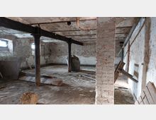 Ein leerer, baufälliger Kellerraum mit freiliegenden Ziegelwänden, einem alten gemauerten Pfeiler, Metallträgern an der Decke und zwei kleinen Fenstern mit Tageslichteinfall. Der Boden besteht aus unebenem Pflaster, und im Raum befinden sich vereinzelte Bauabfälle und Rohre.