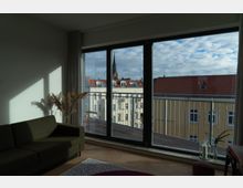 Wohnzimmer mit einem großen bodentiefen Fenster, das auf einen Balkon zeigt. Durch das Fenster sind Wohngebäude mit roten Dächern sichtbar, im Hintergrund ragt der Turm einer Kirche in den bewölkten Himmel.