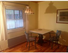 Kleiner Essbereich mit einem quadratischen Tisch und zwei Stühlen aus Holz, in einem Raum mit Holzfußboden und cremefarbenen Wänden. Ein Fenster mit weißen Vorhängen und Häkelgardinen sowie eine hängende Lampe über dem Tisch prägen den Raum; an der Wand hängt ein eingerahmtes Bild.