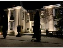 Nachtszene eines großen, beleuchteten Herrenhauses mit heller Fassade, hohen Fenstern und Fensterläden. Im Vordergrund sind schneebedeckte Gehwege, zwei Säulenpflanzen und einige Bäume zu sehen.