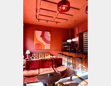 Warm-toned lounge bar interior with red walls, a coffered ceiling and pendant lights, featuring plush velvet seating and a round coffee table in the foreground. A wooden bar counter with tall stools, shelves of bottles, and a wall-mounted chalkboard menu sits along the right side.