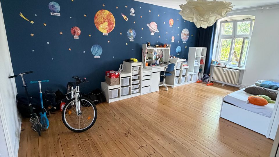 Großes Kinderzimmer mit hellem Holzboden und dunkelblauer Akzentwand, die mit Planeten- und Sternmotiven gestaltet ist. Entlang der Wand stehen weiße Regale und ein Schreibtisch; neben einem großen Fenster mit Heizkörper stehen ein Bett sowie abgestellt ein Fahrrad und ein Roller.