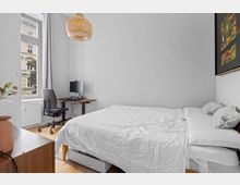 Schlafzimmer mit schlichtem Design: links ein Schreibtisch mit Bürostuhl und Monitor vor einem hohen Fenster, rechts ein großes Bett mit weißer Bettwäsche. An der Decke hängt eine geflochtene Hängelampe, an der Wand ein modernes Kunstwerk.