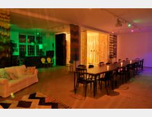 Ein großer Raum mit moderner Einrichtung, beleuchtet in bunten Farben. Links steht ein weißes Sofa vor einer Fensterfront, dahinter befindet sich eine kleine Theke mit gelben Stühlen, rechts ist ein langer Esstisch mit schwarzen Stühlen und Kerzen dekoriert, und farbige Vorhänge trennen Bereiche des Raumes.