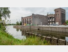 Backstein-Industriegebäude an einem Kanal mit stillgelegter Uferbefestigung; im Vordergrund Wiese und Bewuchs, im Hintergrund Bäume und Strommasten.