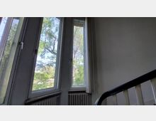 Treppenhaus mit großem Eckfenster, durch das Bäume und ein sonniger Außenbereich sichtbar sind. Rechts ist ein schwarzes Geländer und im unteren Bereich Heizkörper unter den Fenstern erkennbar.