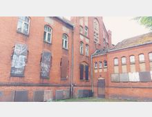 Backsteingebäude mit teilweise vermauerten und vergitterten Fenstern, Graffiti auf mehreren Metallplatten und einem beschädigten Dach; wirkt verlassen und ungepflegt, umgeben von spärlichem Grasbewuchs.