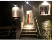 Außenansicht eines Hauseingangs bei Nacht mit eingeschalteter Beleuchtung. Eine steinerne Treppe führt zur halb geöffneten Haustür, daneben ein beleuchtetes Fenster und eine Metallgeländer.