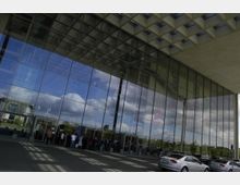 Moderne Glasfassade eines Gebäudes mit reflektierendem Himmel und Wolken; im unteren Bereich sind Menschen versammelt, und mehrere parkende Autos sind auf einem gepflasterten Vorplatz zu sehen.