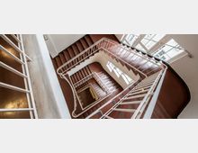 Blick nach unten in ein Treppenhaus mit einem geschwungenen, weißen Geländer und rötlichen Stufen. Das Treppenhaus hat mehrere Etagen und ist von großen Fensterflächen an den Wänden umgeben, die Tageslicht hereinlassen.