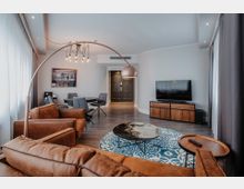 Modernes Wohnzimmer mit braunen Ledersofas, rundem Couchtisch auf einem blauen Teppich mit weißem Muster, einem großen Flachbildfernseher auf einem Holzschrank und einem Esstisch mit vier Stühlen im Hintergrund. Der Raum ist mit einer hohen geschwungenen Stehlampe und großen Fenstern mit halbtransparenten Vorhängen ausgestattet.