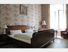 Schlafzimmer mit einem großen Holzbett, weißen Bettdecken und zwei Nachttischen mit Lampen. Die Wände sind mit einer blumigen Tapete in Rosa- und Beigetönen gestaltet, dazu gibt es ein Fenster mit halbtransparenten Vorhängen, zwei Sessel und eine Stehlampe.