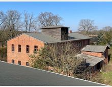 Backsteingebäude im ländlichen Raum mit Satteldach und einem hölzernen Dachaufbau, umgeben von kahlen Bäumen und vereinzelten Nadelbäumen. Neben dem Hauptgebäude stehen kleinere Anbauten mit Wellblechdächern, und im Hintergrund sind Felder und weitere Vegetation zu sehen.