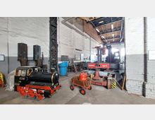 Innenraum einer Werkstatt mit industriellem Charakter. Im Vordergrund steht eine kleine schwarze Spielzeuglokomotive, daneben eine rote Arbeitsplattform mit der Aufschrift „Lokomotor Typ III“; im Hintergrund sind Werkzeuge, Metallkonstruktionen und weiße Wände mit sichtbarem Mauerwerk zu sehen.