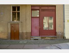 Fassade eines alten Gebäudes mit bräunlicher Putzwand; links eine braune Holztür, daneben ein größerer roter Eingang mit Glasfenster und Tür, teilweise von Graffiti bedeckt. Der Boden besteht aus Kopfsteinpflaster und Gehwegplatten.